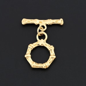 May include: A gold-colored toggle clasp with a bamboo-inspired design. The clasp features a horizontal bar and a hexagonal ring, both with textured detailing. The clasp is designed for jewelry making and is set against a dark background.
