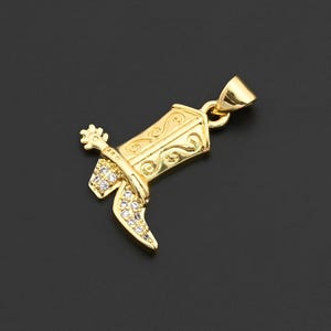 May include: A gold-colored pendant in the shape of a cowboy boot. The boot features intricate scrollwork and is embellished with small, clear gemstones. A spur and a loop for a chain are also visible.