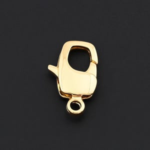 May include: A gold-colored lobster clasp, a jewelry component, is displayed against a dark background. The clasp has a rounded body with a lever and a small loop for attachment. The metal has a shiny, reflective surface.