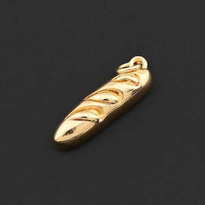 Könnte beinhalten: Ein goldfarbener Anhänger in Form eines Baguettes. Der Anhänger hat eine strukturierte Oberfläche mit detaillierten Rillen und eine kleine Schlaufe oben für eine Kette. Der Hintergrund ist einfarbig schwarz.