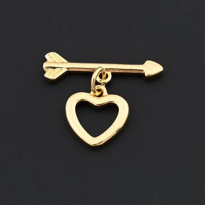 May include: A gold-colored charm featuring an arrow and a heart pendant. The arrow is horizontal with a pointed head and fletching. The heart is a simple, open design. The charm is likely for jewelry making or as a pendant.