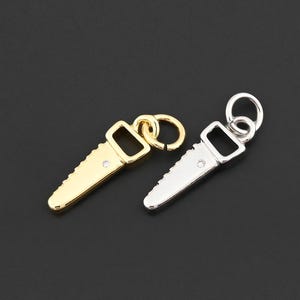 May include: Two small saw-shaped pendants, one gold-colored and one silver-colored, are displayed against a dark background. Each pendant has a small loop at the top for attaching to a chain or bracelet. The saw blades have detailed teeth.