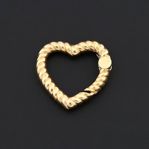 May include: A gold-colored heart-shaped clasp with a twisted rope design. The clasp has a small, round closure on the right side. The heart is set against a dark background.