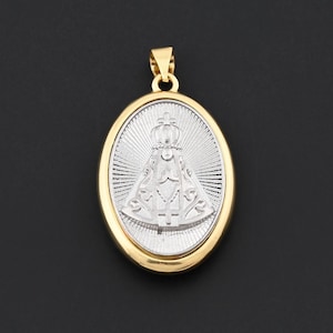May include: Oval-shaped pendant with a gold-colored frame and bail. The pendant features a silver-colored religious figure with a crown and radiating lines. The background is black.