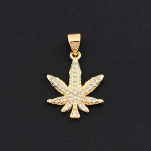 May include: A gold-colored pendant in the shape of a cannabis leaf, encrusted with small, clear gemstones. The pendant has a loop at the top for a chain and is set against a black background.