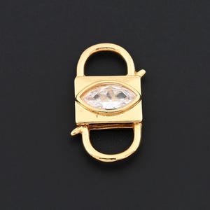 Puede incluir: Un cierre de joyería dorado con un cuerpo rectangular y partes superior e inferior curvas. El cierre presenta una gema transparente con forma de marquesa en el centro. El fondo es negro liso.