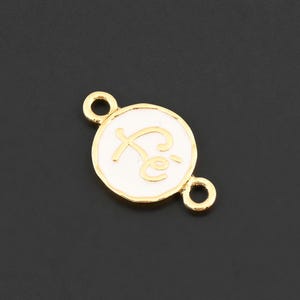 May include: A gold-colored charm with a white enamel center. The charm has the word "Fe" in a cursive font. The charm has two small loops for attaching to a necklace or bracelet. The background is black.
