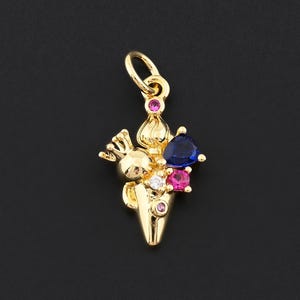 May include: A gold-colored charm featuring a whimsical design. The charm has a cone shape with a small figure and is adorned with blue, pink, and clear gemstones. A small loop is at the top for attachment.