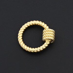 May include: A gold-colored, decorative clasp with a twisted rope design. The clasp features a circular ring and a square-shaped closure, both with a textured, rope-like pattern. The background is a solid black.