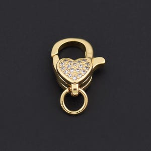 Könnte beinhalten: Ein goldfarbener Verschluss mit einem herzförmigen Mittelteil, der mit kleinen, klaren Edelsteinen besetzt ist. Der Verschluss hat einen kleinen Ring unten und eine gebogene Oberseite, vor einem schwarzen Hintergrund. Dieser Schmuckfund ist für Halsketten und Armbänder geeignet.