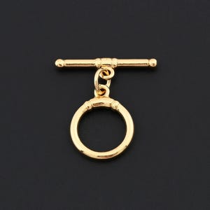 Pode incluir: Um fecho de alternância dourado, apresentando um anel circular e uma barra horizontal com extremidades arredondadas. O fecho é projetado para fazer joias e está sobre um fundo escuro.