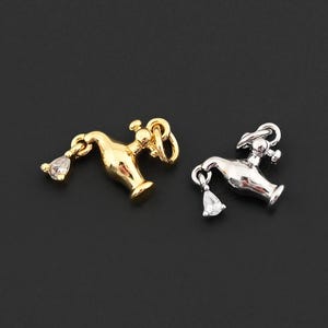 May include: Two small, detailed genie lamp charms. One is gold-colored, the other silver-colored. Each charm has a teardrop-shaped clear gem hanging from the spout. The charms are set against a dark background.