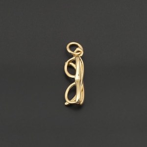 May include: A gold-toned metal charm shaped like a pair of spectacles. The charm features a small circular jump ring at the top for attachment, with a detailed frame design including two circular lenses and a bridge, presented in a vertical orientation.