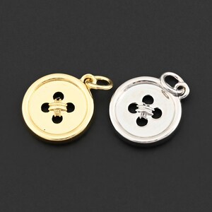 May include: Two button-shaped charms. One is gold-colored, the other is silver-colored. Each charm has a four-petal design in the center and a small loop at the top for attaching to a chain or other jewelry component. The background is black.