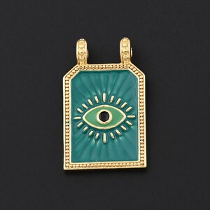 Puede incluir: Colgante dorado con un centro rectangular de esmalte turquesa con un diseño de ojo blanco y verde. El colgante tiene un borde dorado y dos pequeños bucles en la parte superior para una cadena. El fondo es negro.