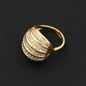 May include: A gold-colored ring with a domed, ribbed design. The dome is encrusted with small, clear gemstones. The ring band is smooth and polished, and the overall design is elegant and eye-catching. The ring is set against a black background.