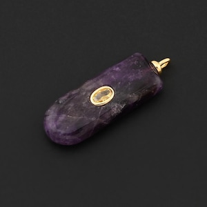 May include: A purple gemstone pendant with a gold-colored bail and a small, oval, gold-colored accent stone. The pendant has a smooth, polished surface and a natural, organic shape. The background is a solid black.
