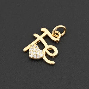 May include: Gold-colored pendant with the word "te" in a cursive script. The letter "t" is stylized, and the letter "e" has a loop. A small heart, encrusted with clear crystals, replaces the dot above the "i".