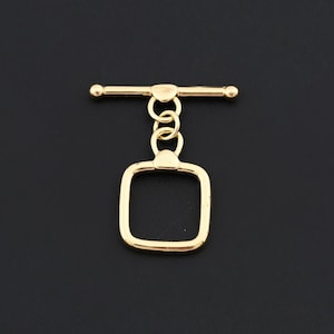 Puede incluir: Un cierre de palanca dorado con un lazo cuadrado y una barra con extremos redondeados, conectados por tres pequeños anillos. El fondo es negro liso.