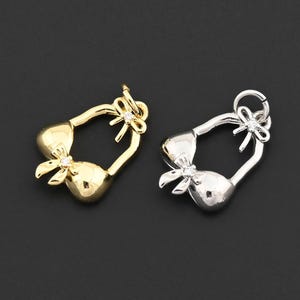 Puede incluir: Dos colgantes metálicos con forma de bikini, uno dorado y otro plateado, sobre un fondo oscuro. Cada colgante presenta un lazo y un diseño delicado, sugiriendo un tema de joyería o artesanía.