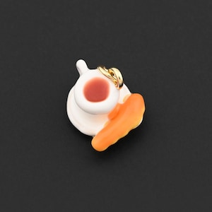 May include: A miniature ceramic teacup and saucer set with a slice of orange pastry. The teacup is white with a red liquid inside. A gold ring is on the saucer. The pastry is orange. The background is black.