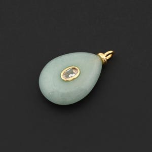May include: A light green teardrop-shaped pendant with a gold-colored bail. The pendant features a small, oval-shaped, clear stone set in a gold-colored bezel in the center. The background is black.