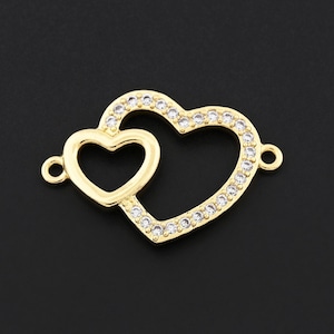 Könnte beinhalten: Ein goldfarbener Anhänger mit zwei verbundenen Herzen. Das größere Herz ist mit kleinen, klaren Kristallen umrandet. Der Anhänger hat kleine Schlaufen an jeder Seite zur Befestigung. Der Hintergrund ist einfarbig schwarz.