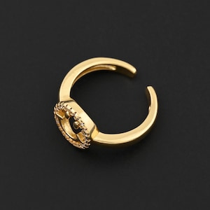 May include: A gold-colored adjustable ring with a circular design. The ring features a small, detailed emblem at the center, surrounded by a textured border. The band is open, allowing for size adjustment. The ring is set against a black background.