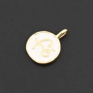 May include: A gold-colored pendant with a white enamel center. The pendant features the word "Fe" in a cursive font. The pendant has a small loop at the top for attaching to a chain or bracelet. The background is black.