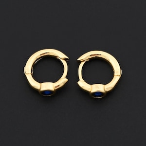 May include: A pair of gold-colored hoop earrings with a small, round, blue gemstone set in the center of each earring. The earrings are displayed against a black background.