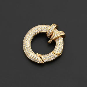 May include: A gold-colored, circular jewelry clasp, encrusted with small, clear gemstones. The clasp has a hinged opening and a decorative band detail. The background is a solid black.