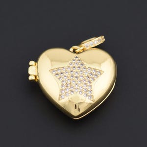 May include: A gold heart-shaped pendant with a star design. The star is set with small, clear gemstones. The pendant has a hinged opening and a small loop at the top for a chain. The background is black.