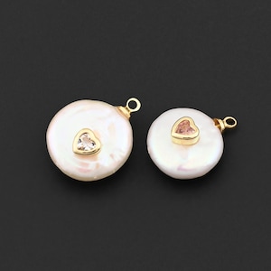 May include: Two white, round pearl pendants with gold-colored accents. Each pendant features a small, heart-shaped gemstone in the center. One gemstone is clear, and the other is pink. Both pendants have a small loop at the top for attaching to a chain.