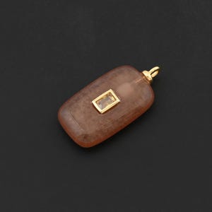 Könnte beinhalten: Ein rechteckiger Anhänger mit einer goldenen Öse und einem kleinen, rechteckigen, goldgerahmten Stein. Der Anhänger hat eine durchscheinende, bräunlich-orange Farbe. Der Hintergrund ist einfarbig schwarz.