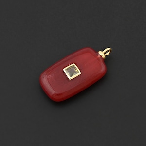 May include: A rectangular red pendant with a gold bail. The pendant features a square, light-coloured stone set in the centre. The background is a solid black.