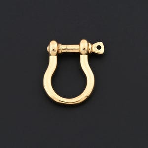 May include: A gold-colored shackle, a U-shaped metal fastener with a pin, is displayed against a black background. The shackle is a bright, shiny gold, and the pin has a small hole. This hardware is often used in jewelry making or for nautical applications.