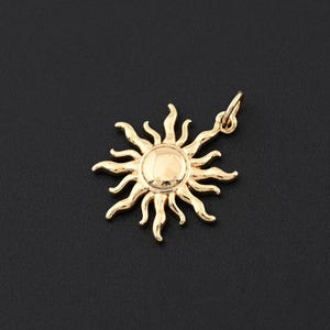 May include: A gold-colored sun pendant with a circular center and radiating rays. The pendant has a small loop at the top for attaching to a chain. The sun design is detailed and shiny, set against a dark background.