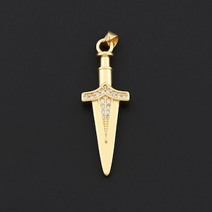 May include: A gold-colored pendant in the shape of a sword. The sword features a crossguard adorned with small, clear gemstones. The pendant has a loop at the top for a chain. The background is a solid black.