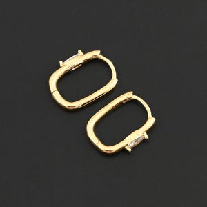 May include: A pair of gold-colored hoop earrings with a rectangular shape. Each earring features a small, clear gemstone. The earrings are displayed on a black background.