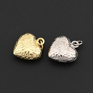 May include: Two heart-shaped pendants. One is gold-colored, the other is silver-colored. Both have a floral pattern and a small loop for attaching to a chain or other jewelry component. The pendants are displayed against a dark background.