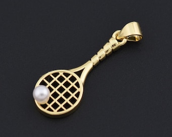 Pode incluir: Um pingente de raquete de tênis dourado com uma pérola no centro da cabeça da raquete. O cabo da raquete tem um design texturizado. O pingente está em um fundo preto.