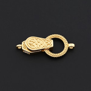 Puede incluir: Un cierre de joyería dorado con un diseño ornamentado. El cierre presenta una caja rectangular con un patrón decorativo y un anillo circular con pequeños detalles de cuentas. El fondo es negro sólido.