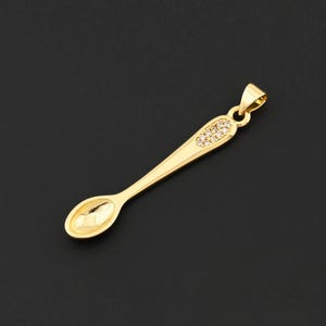 May include: A gold-colored spoon pendant with a small loop at the top for a chain. The spoon's handle is adorned with small, clear crystals. The pendant is set against a dark background.