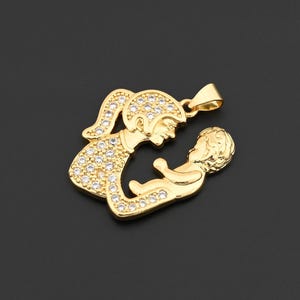 May include: A gold-colored pendant featuring a mother and child design. The mother figure is adorned with small, clear stones, and the baby is held in her arms. The pendant has a loop for a chain.