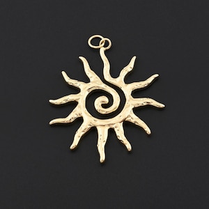 Puede incluir: Un colgante dorado en forma de sol con un centro en espiral y rayos puntiagudos. El colgante tiene un pequeño lazo en la parte superior para colgarlo. El fondo es negro sólido.
