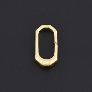 May include: A gold-coloured, geometric clasp with an elongated octagonal shape. The clasp is open, revealing the inner mechanism. The background is a solid black.