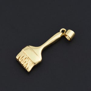 May include: A gold-colored pendant in the shape of a paintbrush. The pendant features a handle, a brush head with bristles, and a loop for attaching to a chain. The pendant is set against a black background.