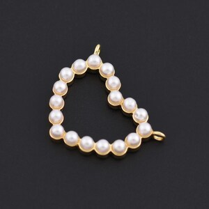 May include: A heart-shaped pendant with a gold-colored frame, filled with small, round, white pearls. The pendant has a loop at the top for attaching to a chain. The background is a solid black.