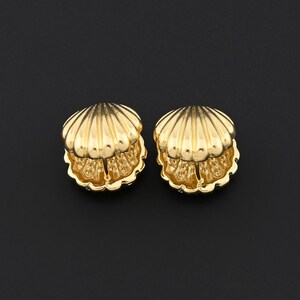 May include: Two gold-colored seashell-shaped earrings. Each earring has a detailed, textured design with a ribbed outer shell and a detailed inner section. The earrings are set against a black background.