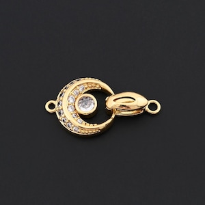 May include: Gold-toned jewelry clasp in a crescent moon shape, embellished with clear crystals. The clasp features a central, larger crystal and smaller crystals outlining the moon shape. The design includes a loop on each end for attachment.